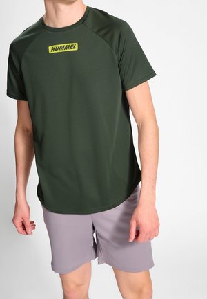 Hombre que lleva una camiseta de manga corta verde oscuro con un logo amarillo de Hummel y pantalones cortos gris claro hasta la rodilla sobre un fondo liso.