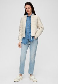 Doudoune beige clair avec un design matelassé, manches côtelées et fermeture éclair à l'avant. Portée sur une chemise en denim bleue et un jean bleu clair.