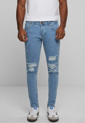 Jeans skinny bleu clair en denim, avec des déchirures distendues au niveau des genoux et des bords effilochés, portés avec des baskets blanches et grises.