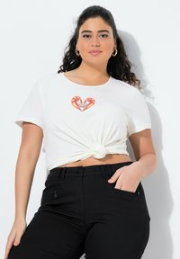 Bílý tričko s krátkým rukávem a uzlem v přední části, s grafikou srdce z oranžových a růžových krevet. V kombinaci s černými vypasovanými kalhotami.