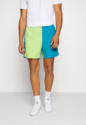 Hombre con pantalones cortos de bloques de color verde y azul, camiseta blanca, calcetines Adidas blancos y zapatillas blancas, de pie contra un fondo liso.