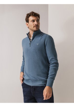 Lichtblauwe trui met ritssluiting aan de hals, geribbelde structuur, lange mouwen en een klein logodetail op de borst, gecombineerd met donkere spijkerbroek.