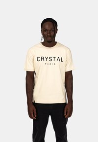 Krémové bavlněné tričko s krátkým rukávem a kulatým výstřihem, na kterém je napsáno "CRYSTAL PARIS" černými blokovými písmeny přes hruď.