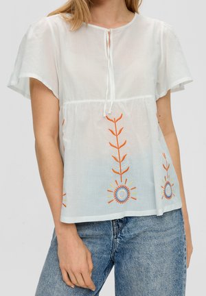 Nainen pukeutuneena valkoiseen lyhythihaiseen paitapuseroon, jossa on oranssin ja sinisen värisiä kukkaompeleita, yhdistettynä sinisiin farkkuihin.