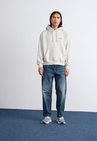A person stands against a plain white wall, wearing a light gray hoodie, blue jeans, and sneakers, with a focused expression.