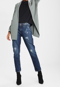 Blazer grigio chiaro su una maglia nera, abbinato a jeans blu strappati. Stivaletti neri alla caviglia completano l'outfit, caratterizzato da una vestibilità rilassata e strappi evidenti.