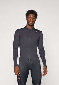 Mann in enganliegendem schwarzen Langarm-Radtrikot und gepolsterten Radhosen, der vor einem schlichten hellen Hintergrund steht.