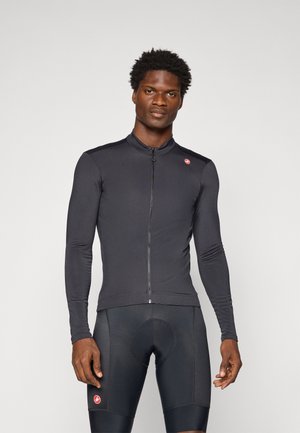 Mann in enganliegendem schwarzen Langarm-Radtrikot und gepolsterten Radhosen, der vor einem schlichten hellen Hintergrund steht.