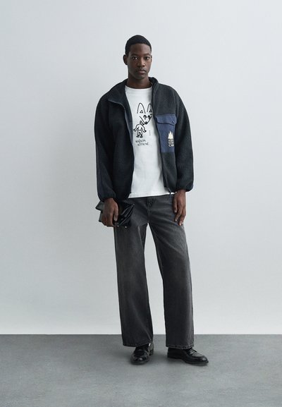 Μαύρο φλις μπουφάν με μπλε λεπτομέρειες πάνω από ένα λευκό μπλουζάκι με γραφικά, συνδυασμένο με γκρί παντελόνι φαρδύ και μαύρα παπούτσια, κρατώντας μια μαύρη τσάντα χεριού.