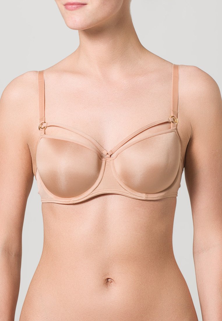 Žena v béžovém podprsenkovém modelu s tenkými nastavovacími ramínky a dekorativními pásky nad košíčky, zachycena od krku po pas.