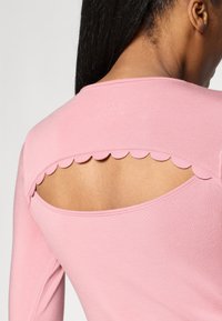 Pink long-sleeve top with a scoop back featuring scalloped edges. Soft fabric with a fitted design, showcasing skin through the cutout.