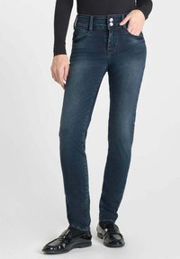Jean skinny bleu foncé en denim, avec une taille haute, fermant par deux boutons et doté de cinq poches. Texture lisse avec un délavage minimal.