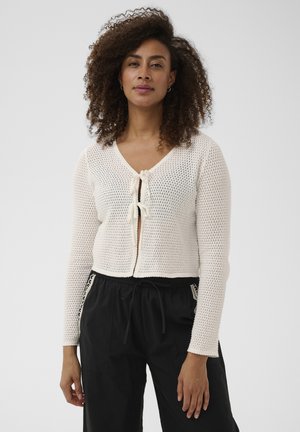 Žena s kučeravými vlasmi oblečená v bielom háčkovanom tričku s dlhým rukávom zviazanom vpredu a v čiernych voľných nohaviciach so sťahovacím pásom.