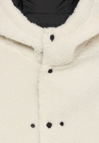 Krémový fleece kabát s kapucí, černými patentkami a měkkým, texturovaným povrchem. Podšívka z tmavého materiálu.