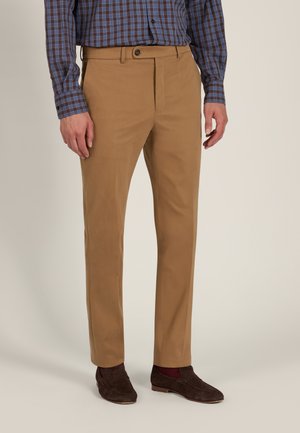 Man met beige pantalon, bruine loafers, bordeauxrode sokken en een blauwbruine geruite blouse met lange mouwen tegen een effen achtergrond.