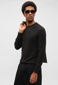 Černý texturovaný svetr s kulatým výstřihem, v kombinaci s černými kalhotami s vysokým pasem. Modelka nosí tmavé sluneční brýle a drží hnědý předmět.