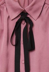 Blusa rosa chiaro con colletto tondo, fiocco in raso nero e bottoni coordinati. Tessuto morbido con pieghe verticali sulle spalle.