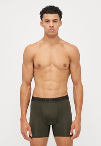 Boxer briefs em tecido verde-oliva com uma textura suave. A cintura possui uma faixa preta com logotipo. O modelo está em pé, exibindo o ajuste.