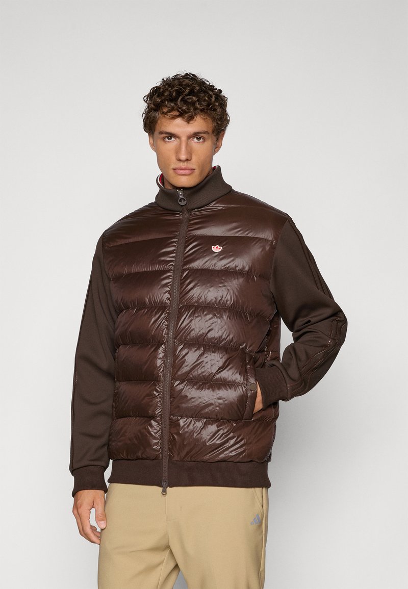 Smeđi prsluk s perjem s sjajnijom, kviltanom gornjom sekcijom i pletenim smešnim rukavima. Ima visok ovratnik i mali logo na grudima.