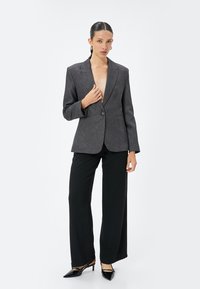 Grijze op maat gemaakte blazer met scherpe revers, enkele knoopsluiting en gestructureerde schouders, gecombineerd met zwarte brede pijpen en puntige schoenen.