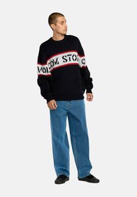 Suéter de punto azul marino con rayas horizontales rojas y blancas; presenta un diseño de texto en negrita. Combinado con jeans azules holgados y zapatos negros.