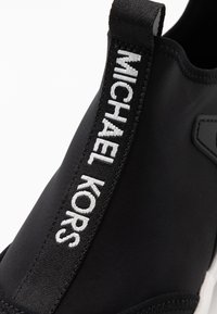 Czarne sneakersy z elastyczną cholewką, z wyraźnym białym logo "MICHAEL KORS" po boku. Gruba biała podeszwa i gładka faktura.