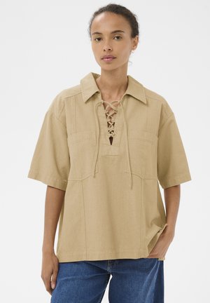 Mujer con camisa beige de manga corta con cordones en el frente y cuello, combinada con jeans azules, sobre un fondo blanco liso.