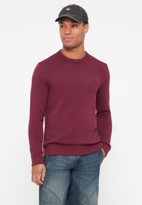 Burgundski pleteni pulover z rebrastim okroglim izrezom in dolgimi rokavi. Nosite ga z denim kratkimi hlačami. Sivi klobuk z logotipom na sprednji strani.