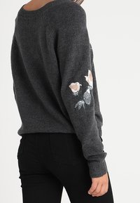 Kobieta w ciemnoszarym swetrze z jasnoróżowym haftem kwiatu na rękawie, w połączeniu z czarnymi spodniami, stojąca tyłem do aparatu.