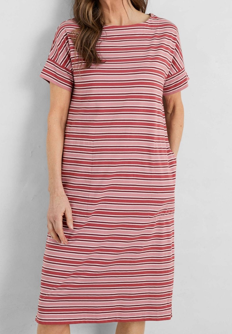 Femme portant une robe courte jusqu'aux genoux à manches courtes avec des rayures horizontales roses et rouges, debout avec une main dans la poche.