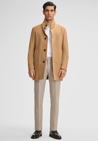 Cappotto in misto cammello con colletto alto, cinque bottoni neri e due tasche laterali, abbinato a pantaloni beige chiaro su misura e scarpe nere.