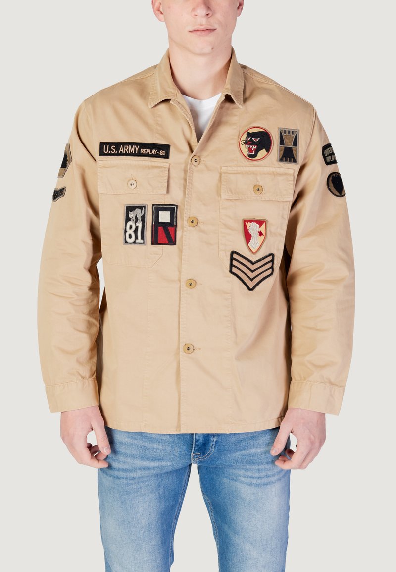 Beige arbejdsjakke med flere broderede mærker; har knaplukning fortil, to brystlommer, lange ærmer og skjortekrave.