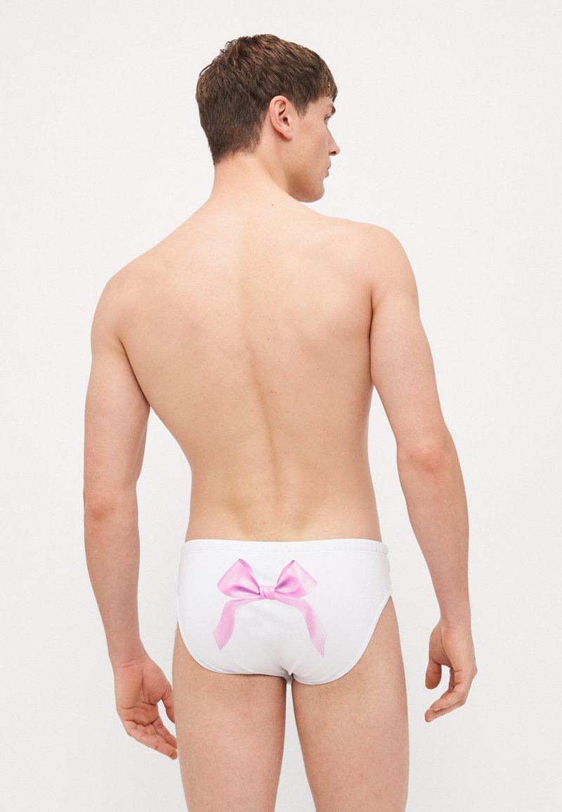 Les slip de bain blancs présentent un grand nœud rose à l'arrière. Le tissu semble lisse avec un ajustement serré, conçu pour le confort.