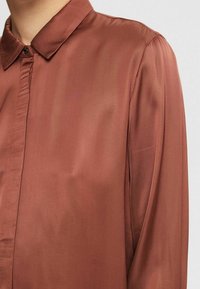 Close-up van een persoon die een roestkleurige satijnen blouse draagt met een puntige kraag en verborgen voorknoopjes.