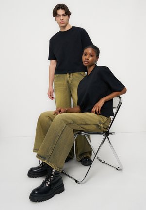 Mann som står og kvinne som sitter på en metallstol, begge iført svarte topper og gulaktige vide bukser med svarte støvler, foran hvit bakgrunn.