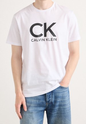 Mann trägt weißes Calvin Klein T-Shirt mit schwarzem CK-Logo und blauen Jeans, steht mit einer Hand in der Tasche und der anderen seitlich.