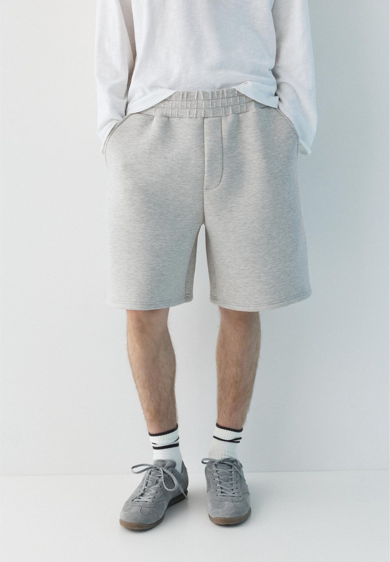 Homme portant un short gris clair à taille élastique, une chemise blanche à manches longues, des chaussettes blanches aux chevilles avec des rayures noires et des baskets grises, debout contre un mur blanc.