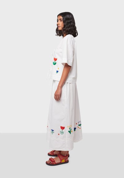 Witte katoenen top met bloemembroidery in rood, groen en blauw. Gecombineerd met een bijpassende witte rok versierd met vergelijkbare patronen. Rode sandalen.