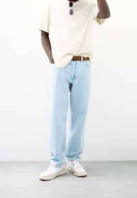 Jeans de color azul claro con corte relajado, combinados con una camiseta color crema. Realzados con un cinturón marrón y zapatillas blancas. Sin patrones visibles.