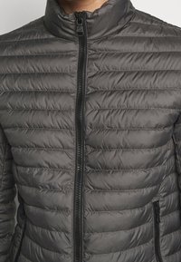 Grå pufferjacka med horisontellt sydda paneler, hög krage, framzipper och fickor på sidan med svarta dragkedjor.