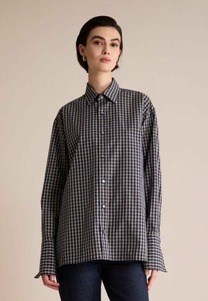 Femme portant une chemise à carreaux ample à manches longues bleu marine et beige avec un jean foncé sur un fond beige uni.