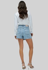 Shorts de mezclilla en azul claro con cintura alta, detalles desgastados y bolsillos traseros, combinados con una camisa blanca de manga larga y tacones plateados.