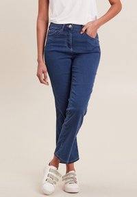 Jeans in denim blu scuro con un taglio aderente, leggermente svasati alla caviglia, cinque tasche e chiusura classica con bottone. Indossati con sneakers bianche.