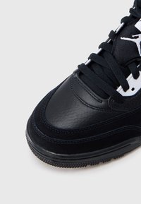 Μαύρο δερμάτινο αθλητικό παπούτσι με λείο φινίρισμα, με ανάγλυφο πάνελ, μαύρα κορδόνια και λευκό λογότυπο σε μαύρο φόντο.