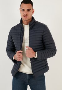 Giacca di piumino blu navy con cuciture orizzontali, colletto alto e chiusura con zip. Indossata sopra un maglione di maglia crema e jeans scuri.