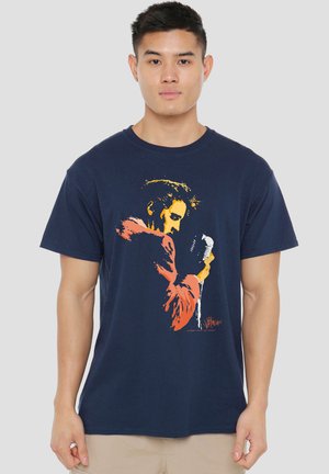 Junger Mann trägt ein marineblaues T-Shirt mit einer orange-gelben Grafik eines Sängers, der ein Vintage-Mikrofon hält.