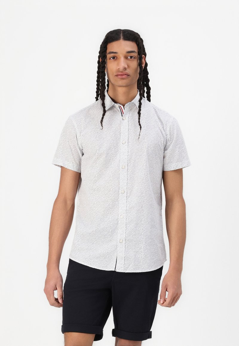 Camicia a maniche corte con bottoni di colore bianco con un motivo a pois neri, dotata di colletto con accenti rossi e blu. Indossata con pantaloni corti blu navy.