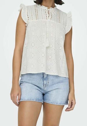 Vrouw die een mouwloze witte eyelet blouse met gerimpelde schouders en lichtblauwe denim shorts draagt, staand met ontspannen armen.
