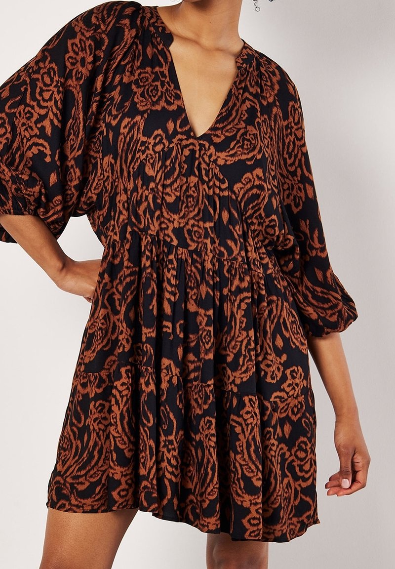 Femme portant une robe noire courte et ample avec des motifs floraux orange rouille et des manches bouffantes trois-quarts, posant avec une main sur la hanche.