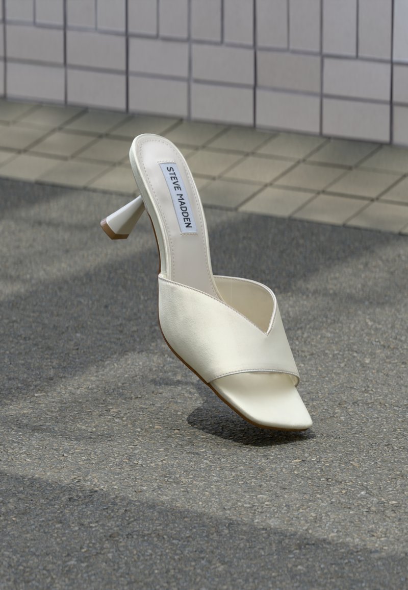 Mule blanche Steve Madden à bout ouvert avec talon bloc reposant sur un asphalt gris près d'un mur carrelé en arrière-plan.